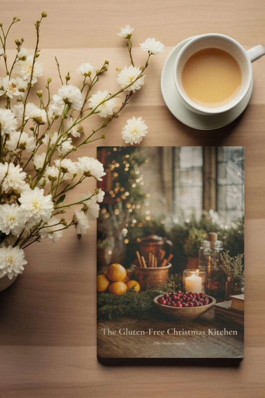 A cosy overhead scene with a cup of tea and soft white flowers arranged beside a copy of The Gluten-Free Christmas Kitchen cookbook.
