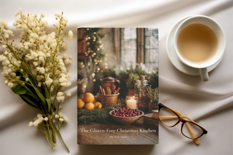 A cosy overhead scene with a cup of tea and soft white flowers arranged beside a copy of The Gluten-Free Christmas Kitchen cookbook.
